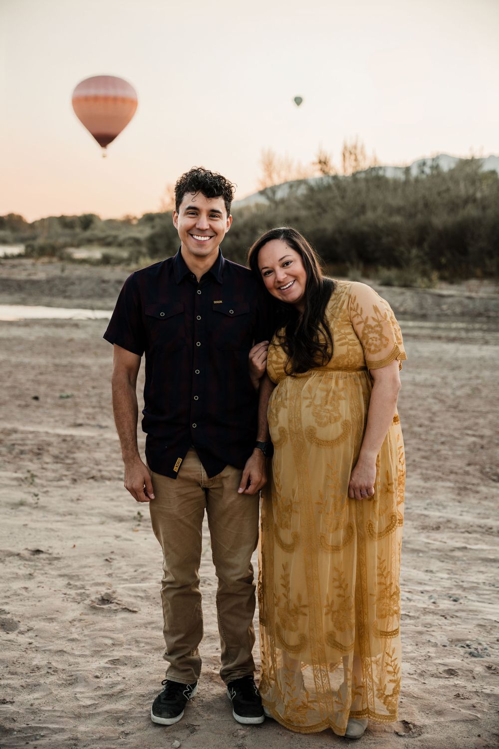 A couple poses with hot air balloons floating in the desert background at sunset.