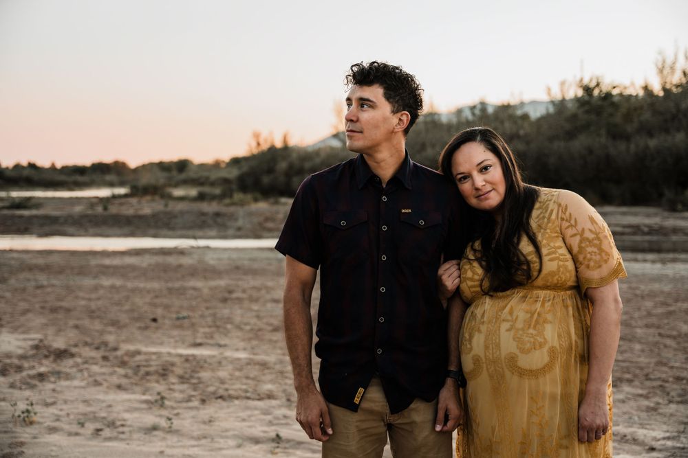 Couple poses together during sunset maternity session near water in desert landscape.