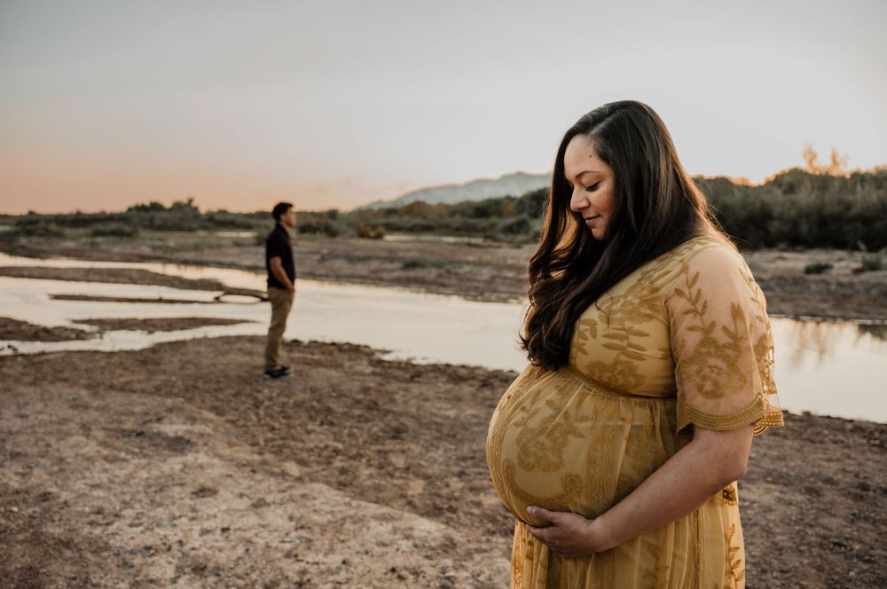Maternity photo shoot at sunset by a lake with woman in yellow lace dress and figure in distance.