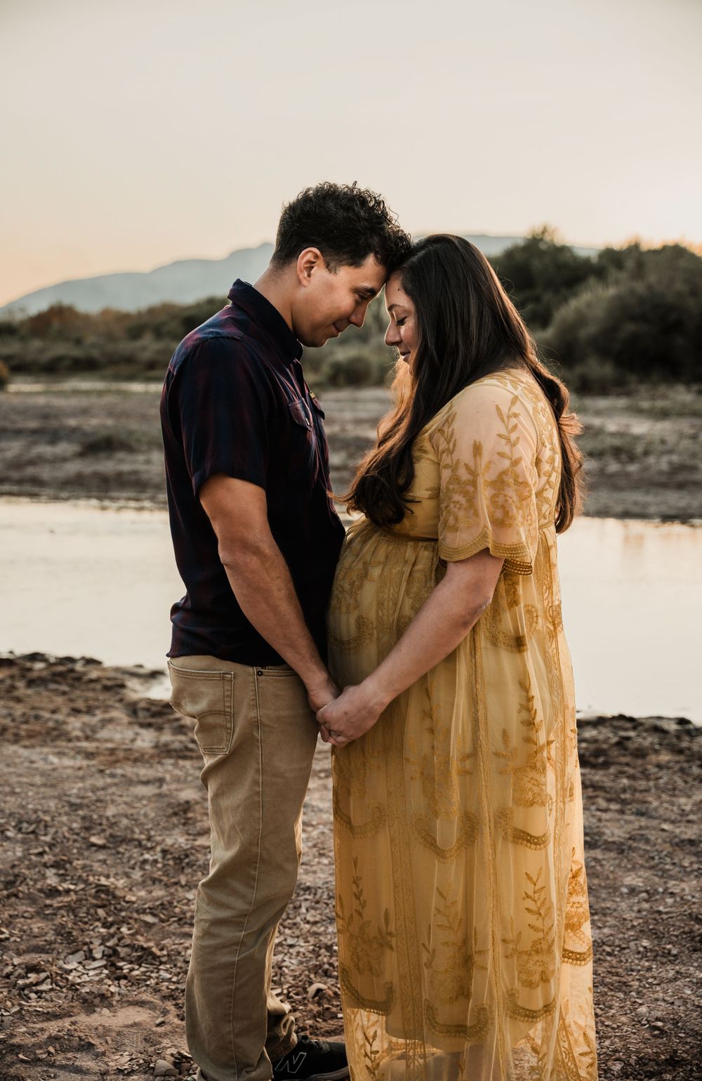 Intimate moment between expectant couple in desert setting with water reflection at sunset.