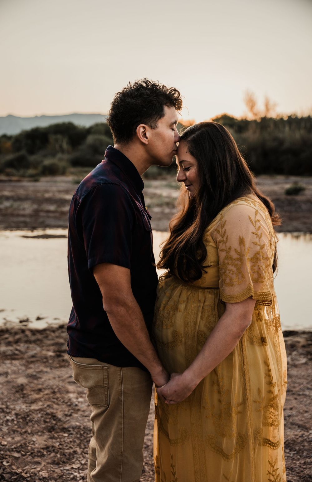 Romantic maternity photo of couple in close embrace by desert lake at golden hour.