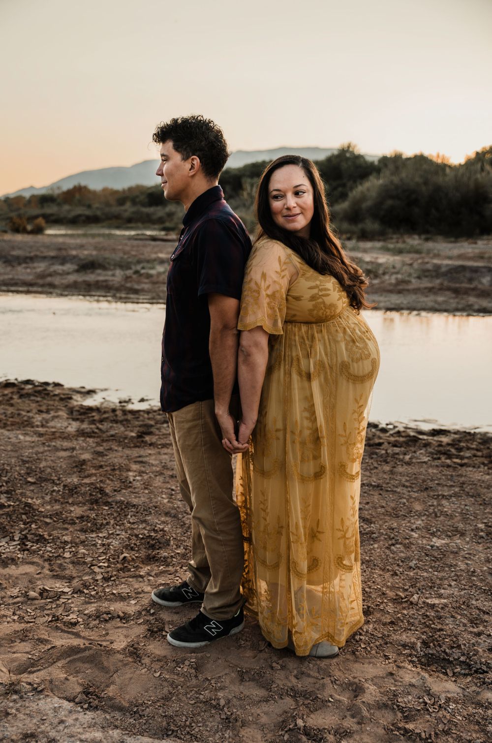 Couple holds hands while standing back-to-back during sunset maternity shoot by lake.