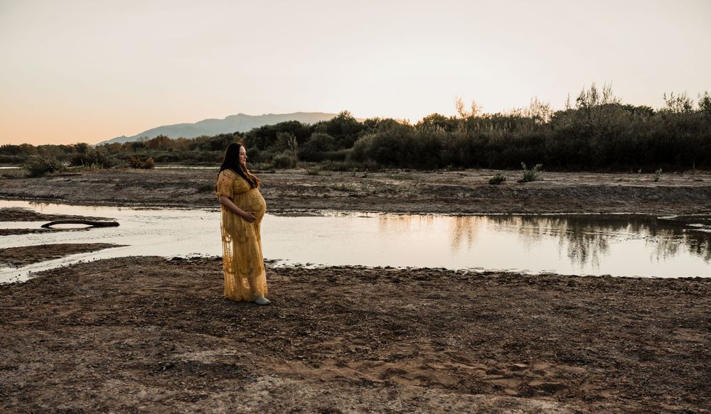 Solitary figure in flowing dress stands by desert lake at sunset.