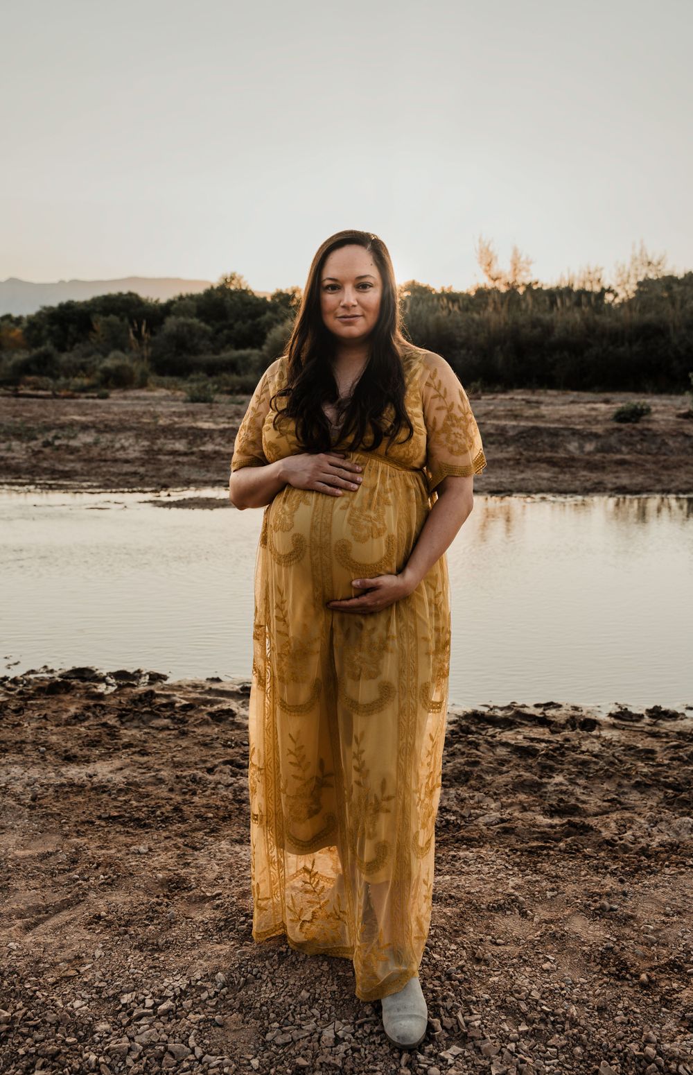 Expectant mother in yellow lace dress poses by lake during golden hour photoshoot.