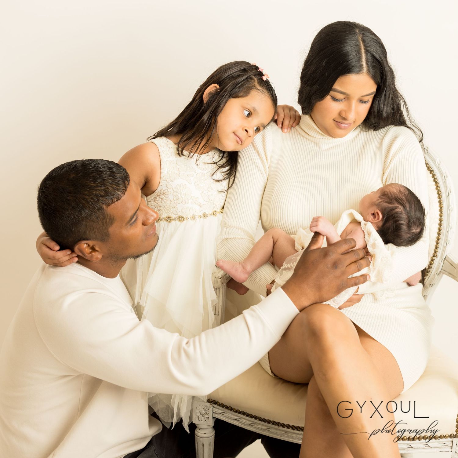 Family in white outfits sharing tender moment with their newborn baby in studio setting.