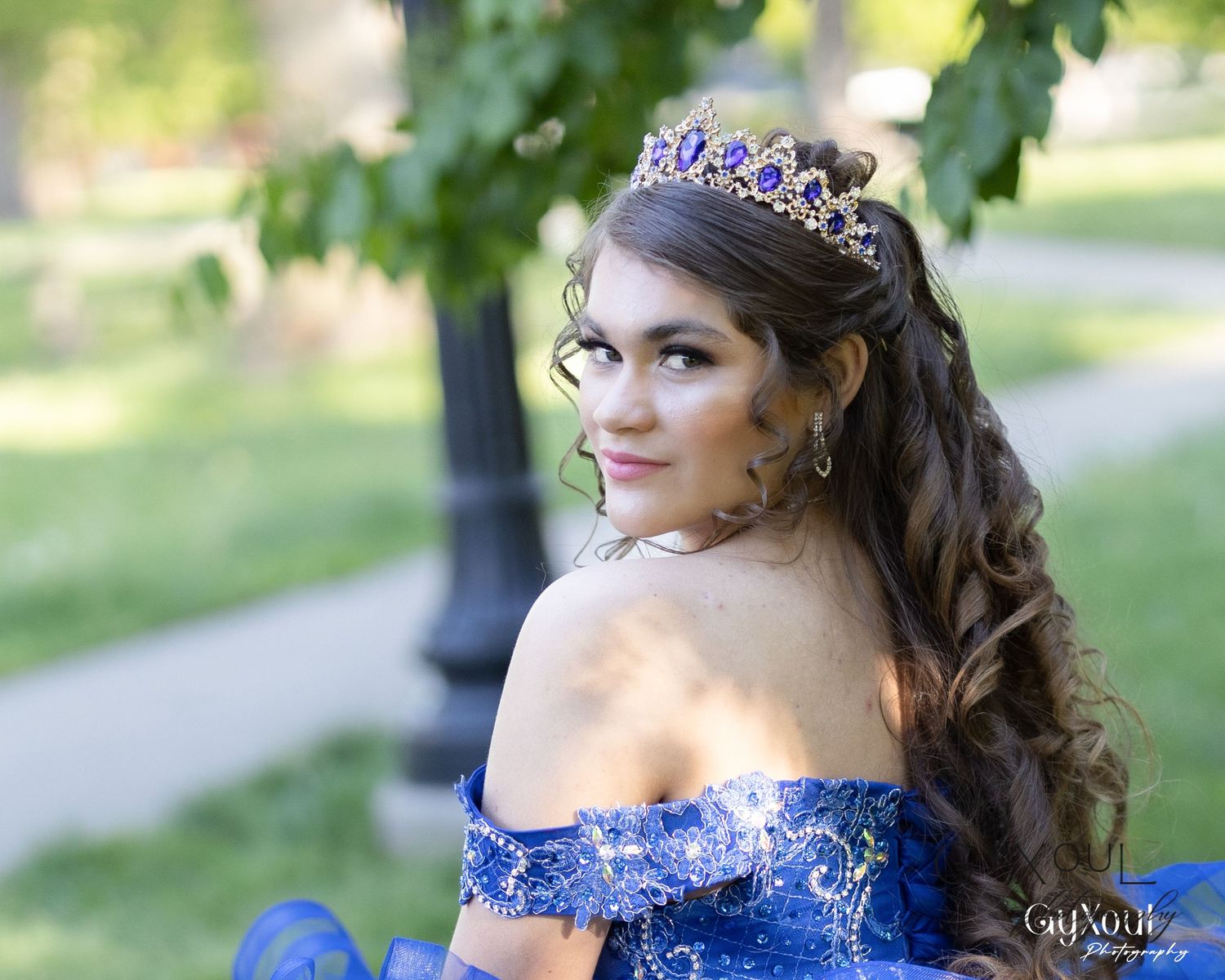 Elegant royal blue off-shoulder gown with sparkly tiara and curled hairstyle in outdoor garden setting.