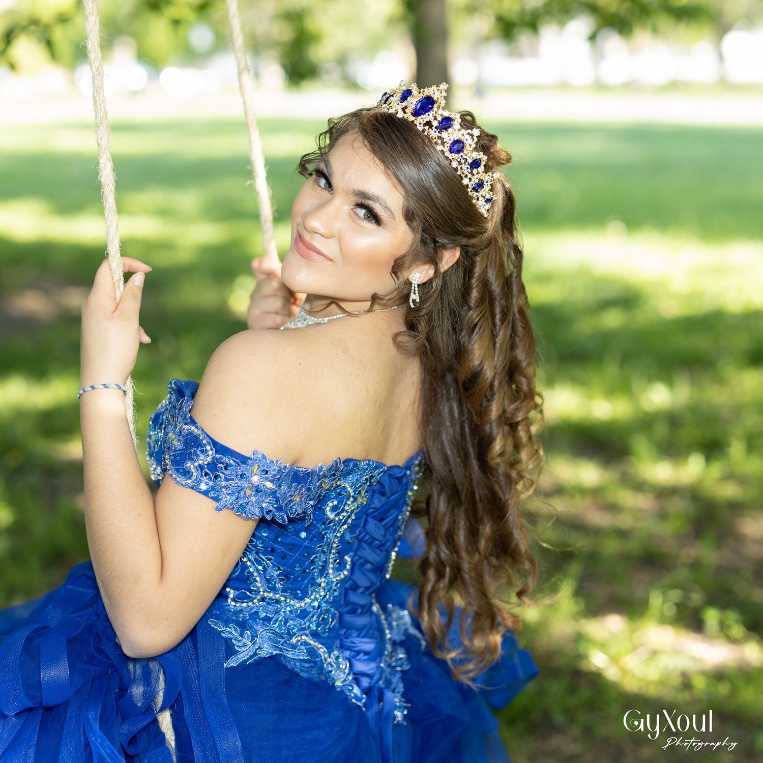 Stunning royal blue quinceañera dress with off-shoulder design and sparkly tiara photographed in a sunny garden setting.