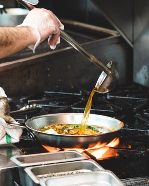 Chef pouring golden sauce into a sizzling pan with herbs over an open flame in a restaurant kitchen.