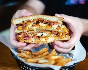 Hands holding a toasted sandwich filled with buffalo chicken, bacon, and cheese over a basket of fries.