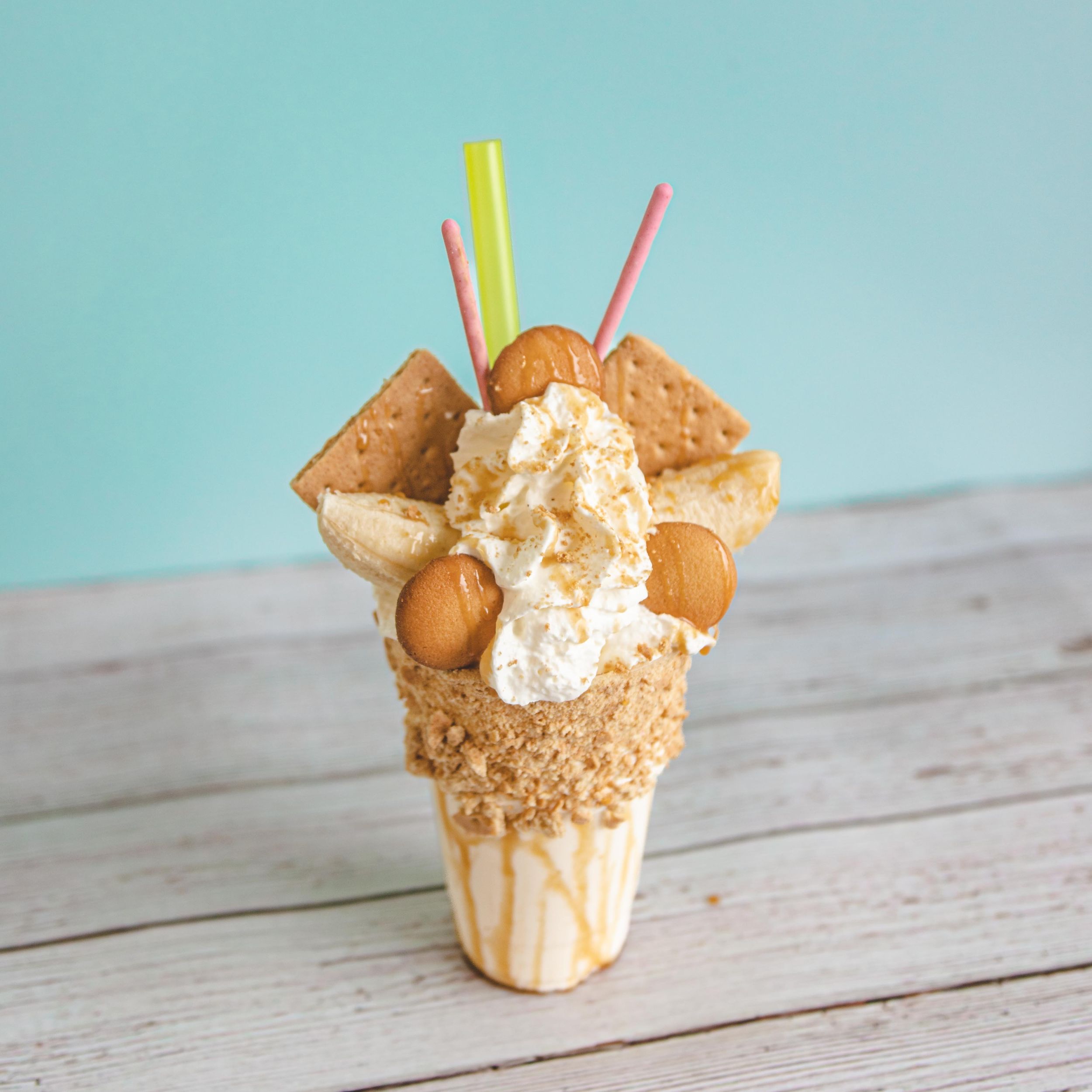 Festive banana pudding milkshake with graham crackers, vanilla wafers, and colorful straws in a caramel-drizzled cone.