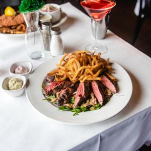 Sliced medium-rare steak with crispy fries on a white plate, served with a pink cocktail and sides.