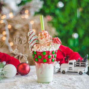 Festive Christmas freakshake with green straw, M&Ms, whipped cream, and holiday toppings in snowy setting.