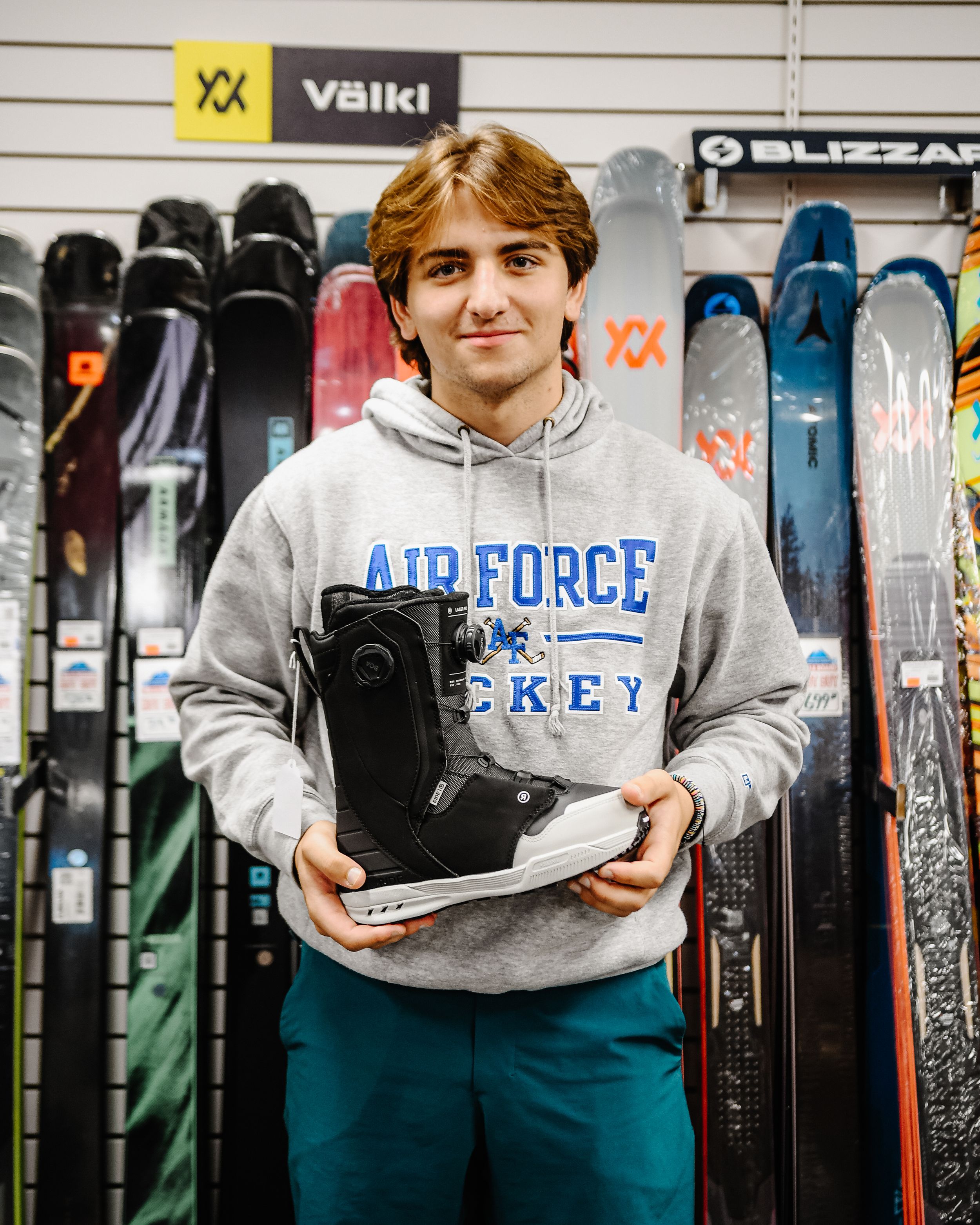 Young man in Air Force Hockey hoodie holding snowboard boots in a ski and snowboard shop.