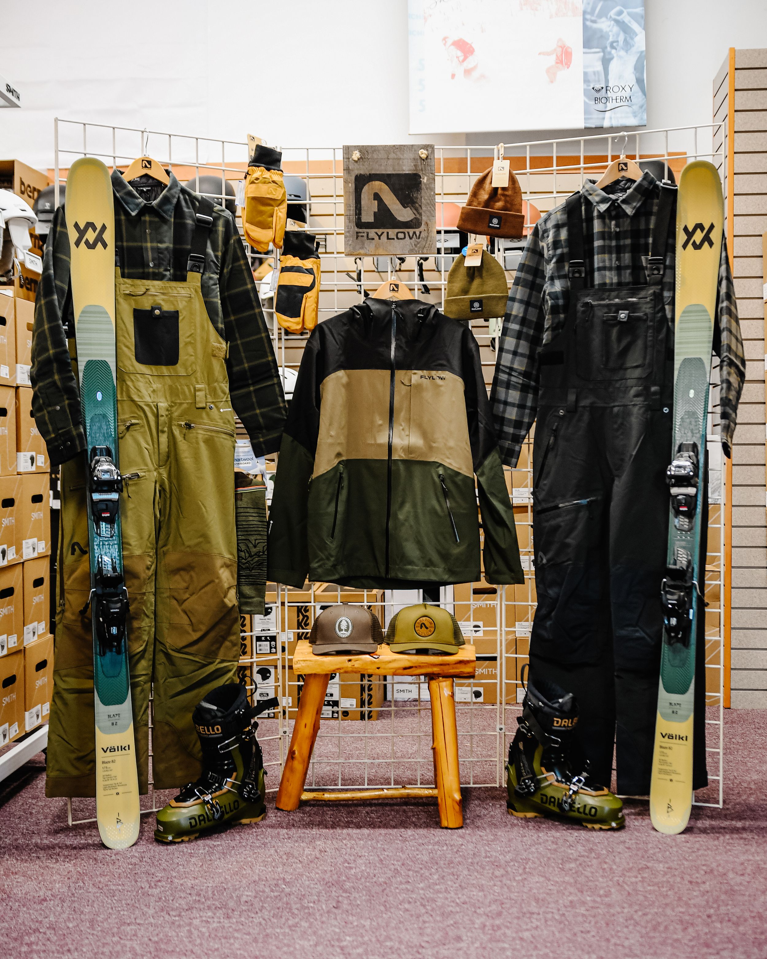 Lylow ski outerwear display featuring bibs, jacket, skis, and boots arranged in a retail ski shop.