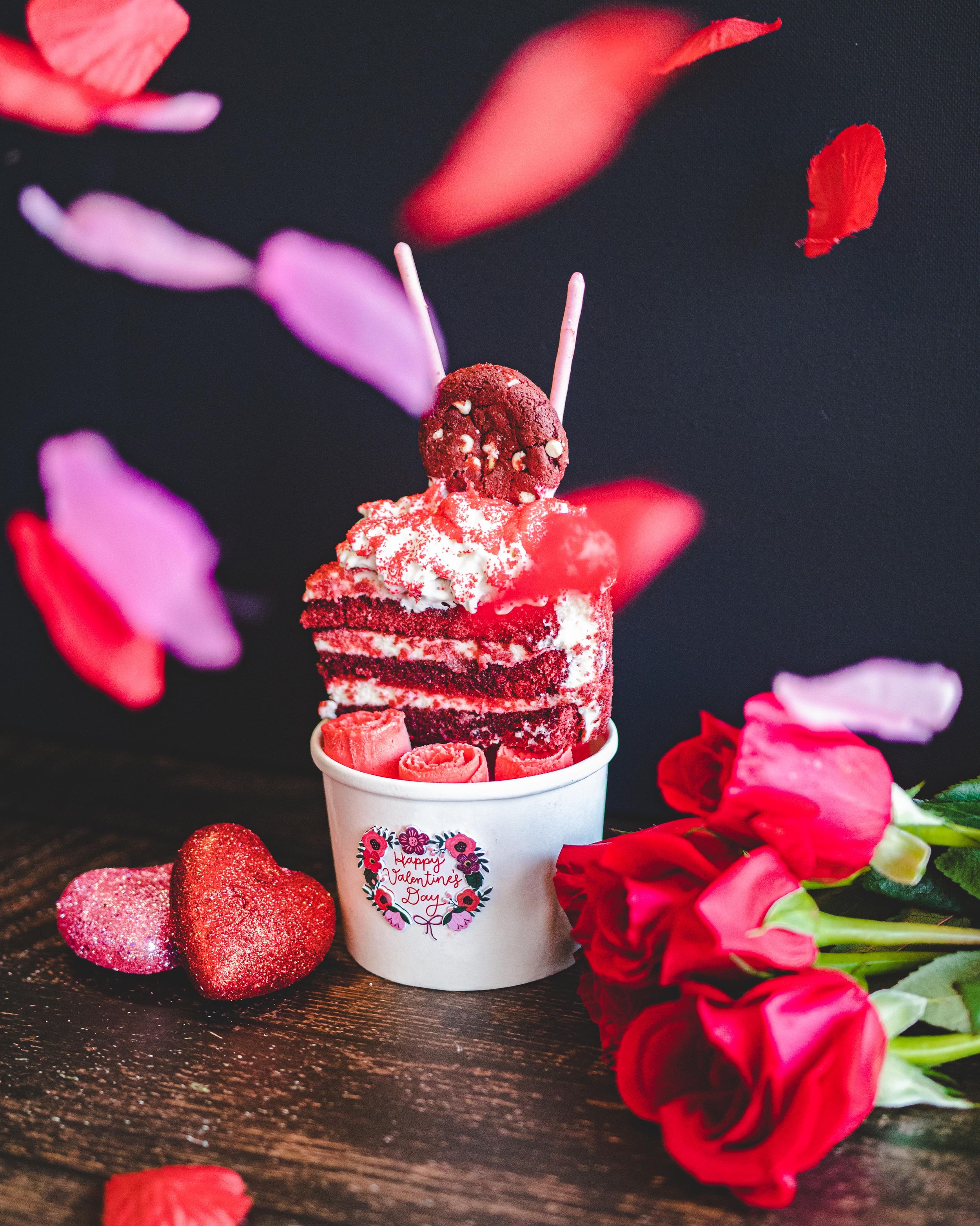 Red velvet cake freakshake in a white cup topped with cake layers and lollipop, surrounded by roses and petals.