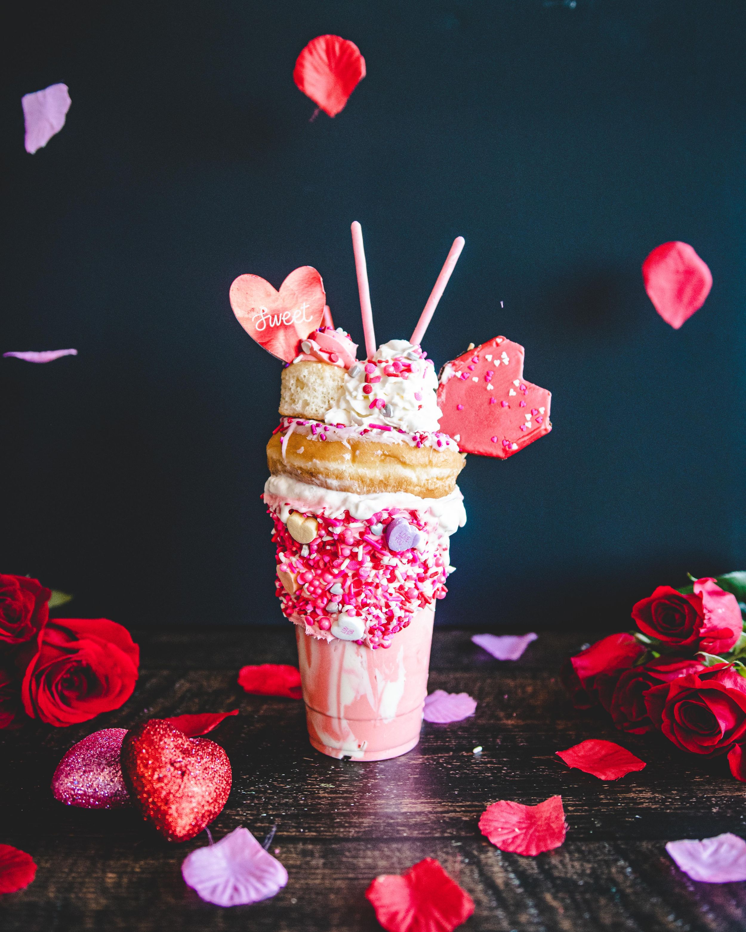 Extravagant Valentine's Day freakshake with pink sprinkles, donuts, heart decorations, and rose petals falling around it.
