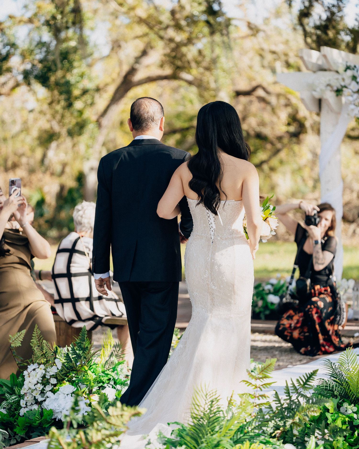 Bride in fitted lace gown walks down outdoor aisle with escort through lush greenery and sunlit oak trees.