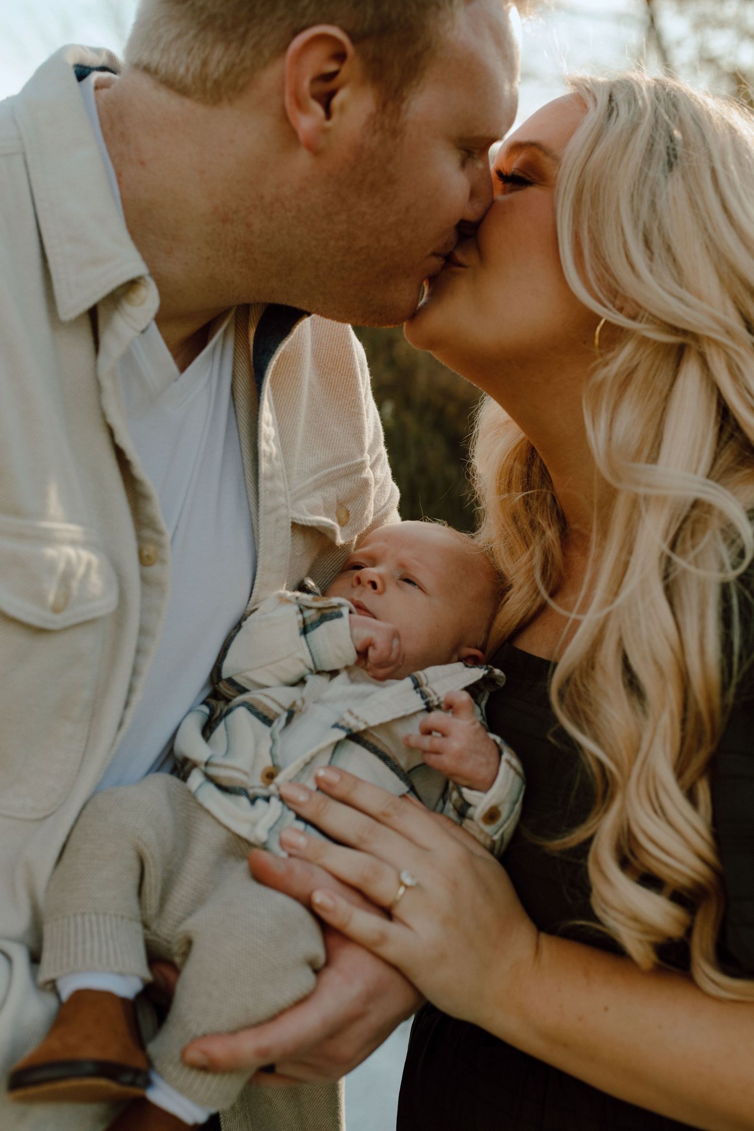 A tender family moment as parents share a kiss while holding their newborn baby in warm sunlight.