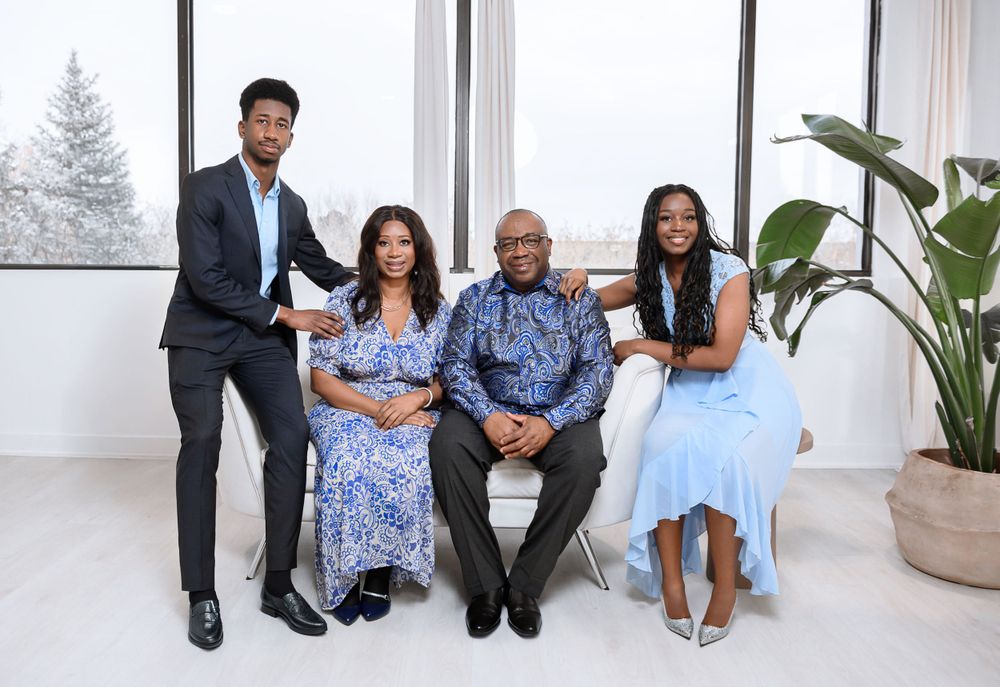 A professional family group portrait in formal attire sitting together in a bright modern interior with large windows and plants.