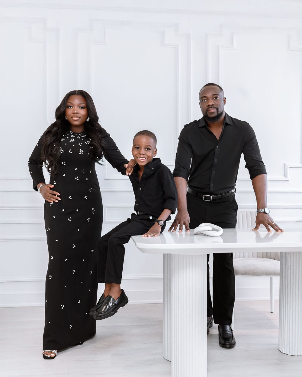 A family of three poses together in coordinated black outfits against a white backdrop with modern white furniture.
