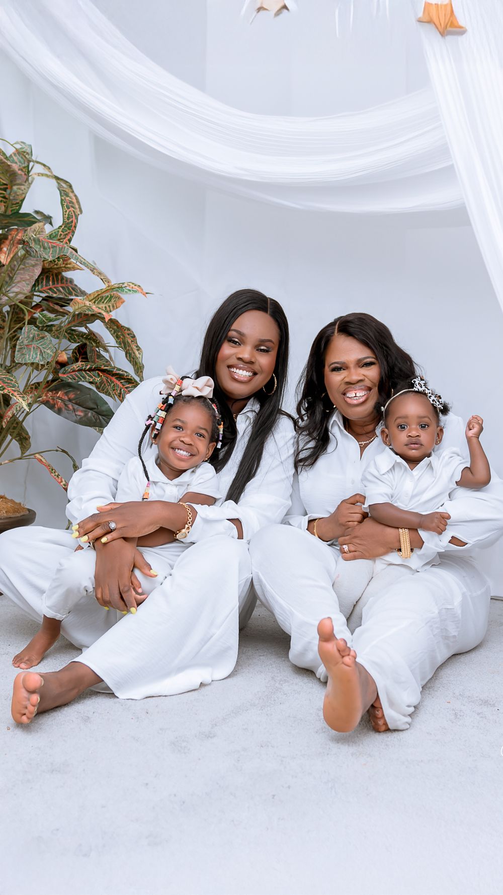 A family portrait featuring matching white outfits sits together on the floor under draped fabric with decorative plants.