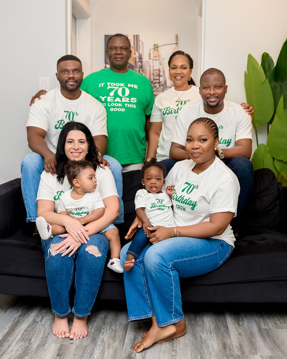 Group wearing matching green and white t-shirts pose together on a gray couch in a modern living room setting.