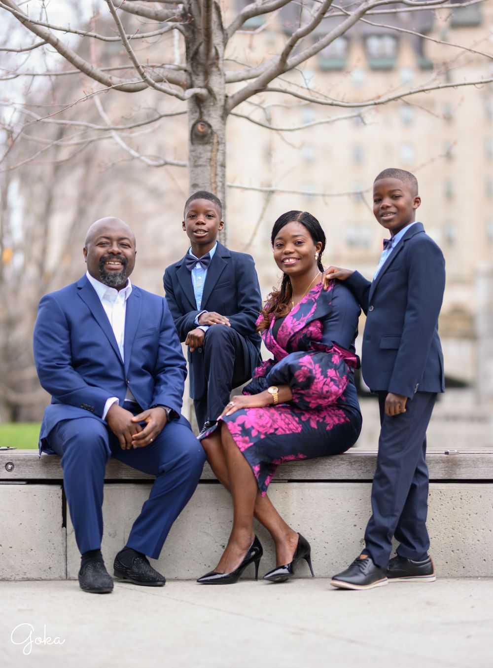 A family of four poses together outdoors in coordinated formal attire with navy suits and a floral dress.