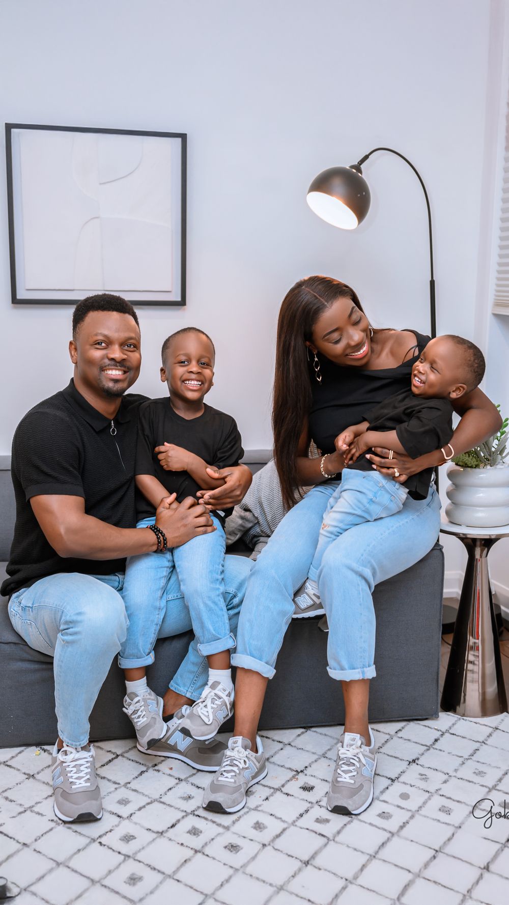 Family wearing matching gray sneakers and blue jeans sits together laughing on a couch in a modern living room setting.