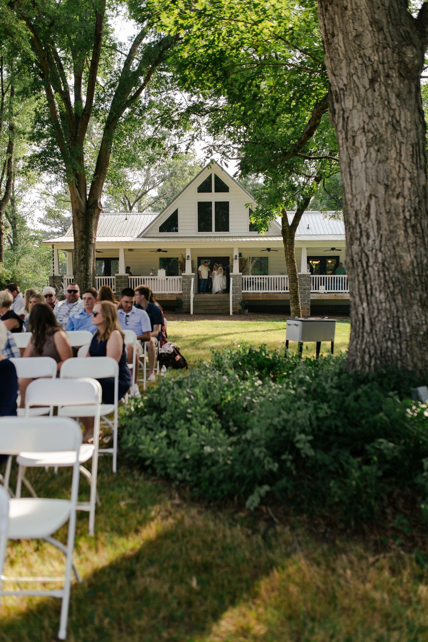 White chairs set up for an outdoor wedding ceremony in front of a charming white farmhouse with a wraparound porch.