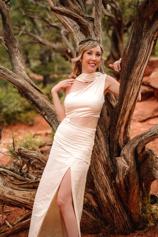 Person in white dress poses elegantly against twisted tree trunk in Sedona desert setting.