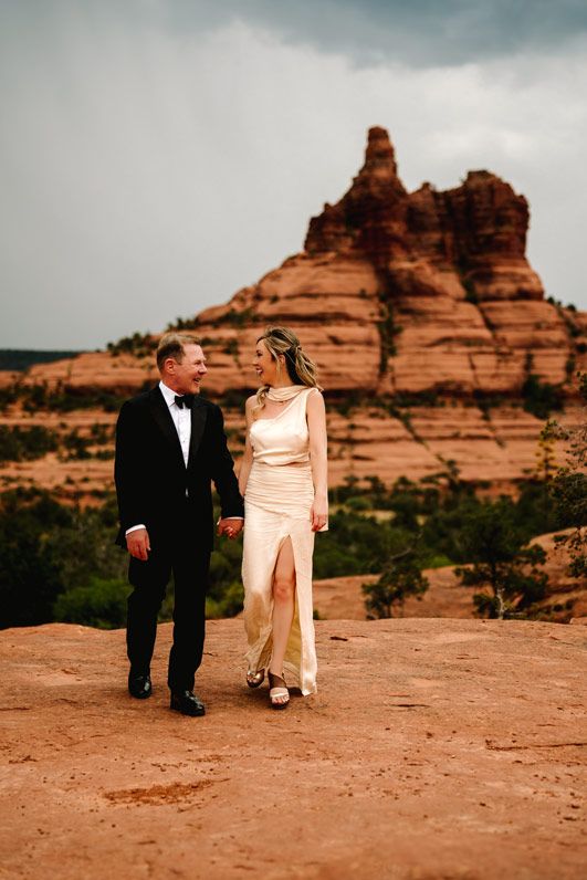 Elegant couple in formal wear stands before towering red rock formations in Sedona.