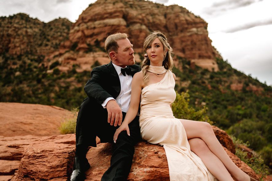 Couple in formal wear sits intimately on Sedona red rock overlook with mountain backdrop.