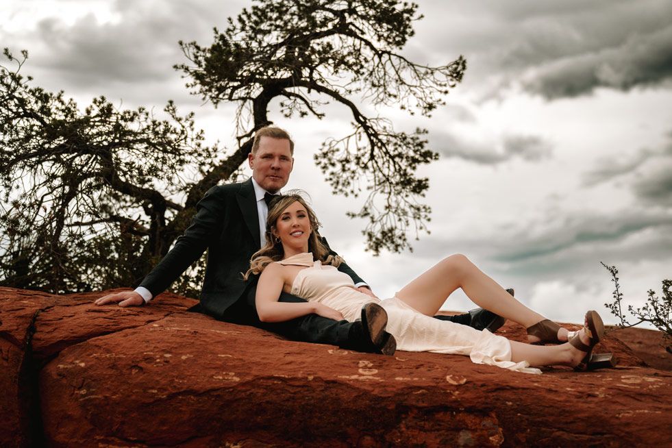 Couple in formal wear poses on Sedona red rock formation under dramatic cloudy sky.
