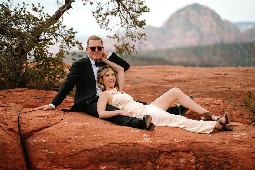 Couple in wedding attire relaxes on red rocks in Sedona with mountain views in the background.