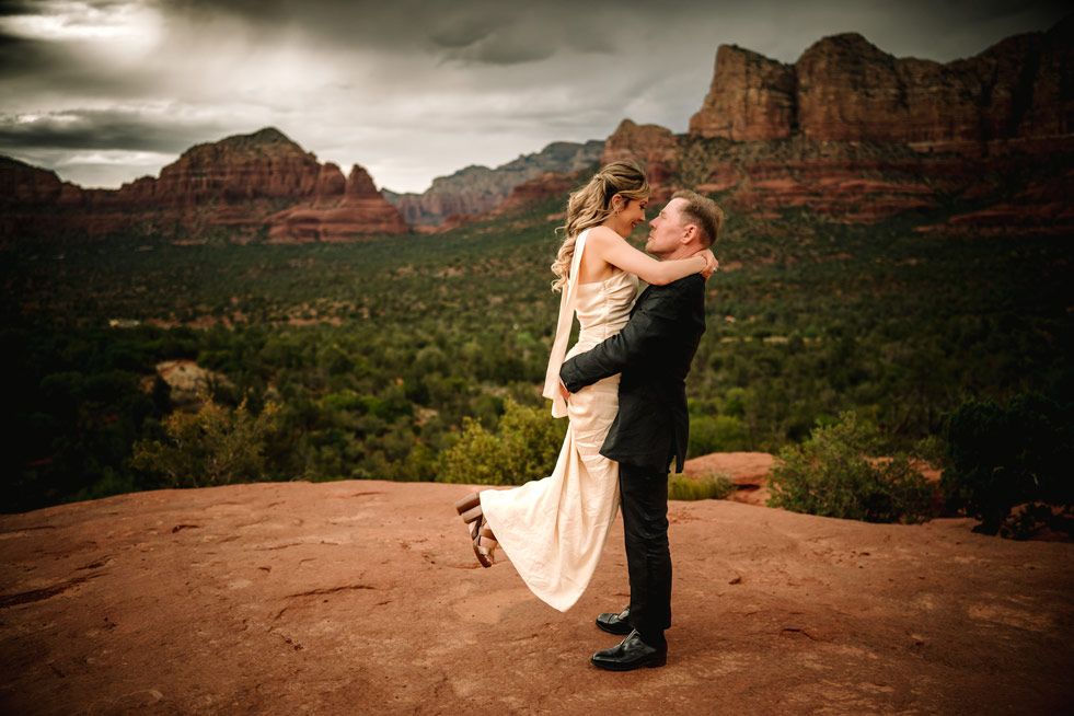 Romantic embrace on Sedona red rock overlook with dramatic desert valley and cliffs in background.