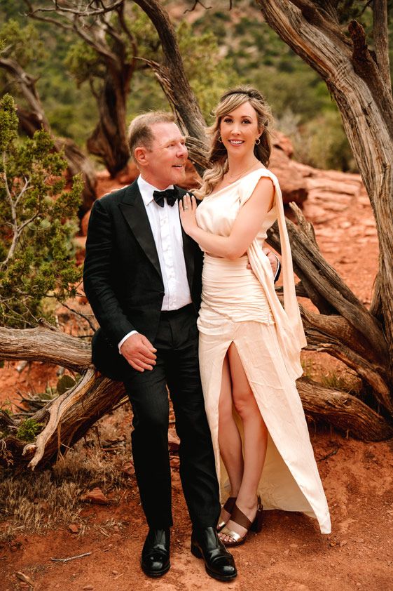 Couple in formal wear poses by twisted tree trunk on desert Sedona red rocks.