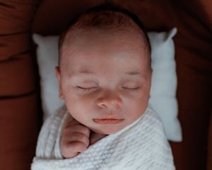 A peaceful sleeping newborn baby swaddled in a white blanket rests on a dark brown pillow.