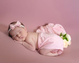 Sleeping newborn baby wearing pink headband posed on pink background with flower.
