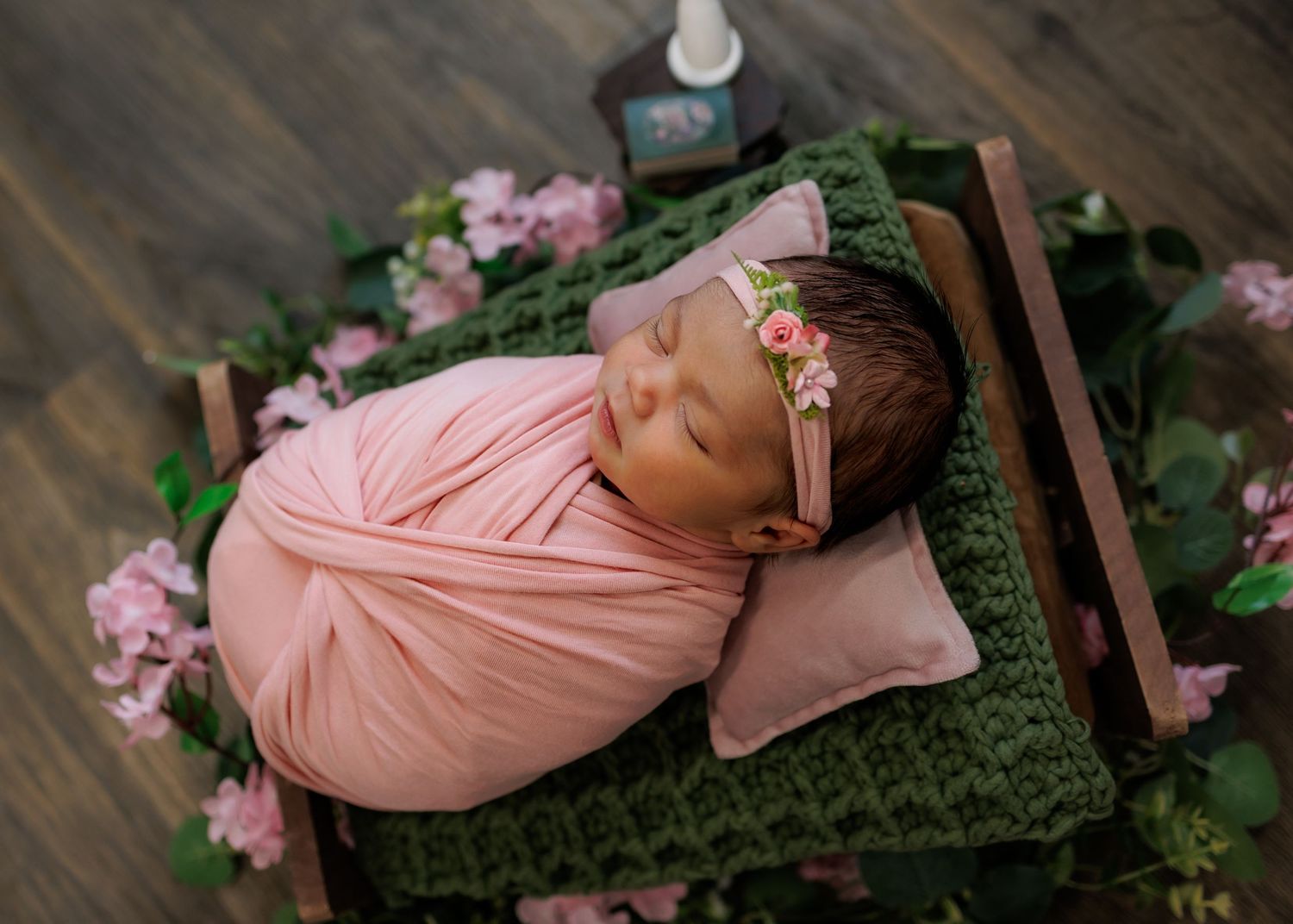 Newborn baby swaddled in pink wrap sleeping on tiny wooden bed with green crochet blanket and pink floral headband.