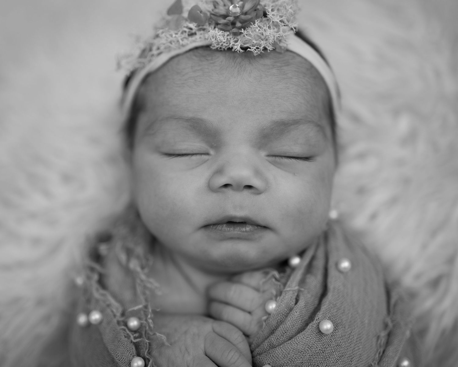 Black and white newborn portrait of a sleeping baby girl wearing a floral headband and pearl-adorned wrap.