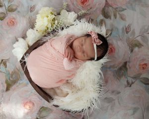 Newborn dressed in pink romper sleeping in wicker basket with white feathers on floral background.