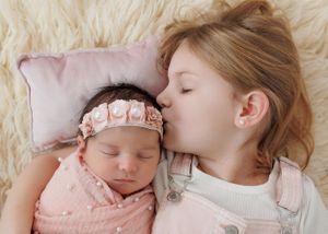 A sweet tender moment of sisters lying close together on a fluffy white rug with the younger wearing a pink floral headband.