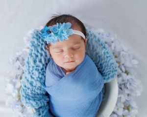 A sleeping infant wrapped in soft blue fabric lies in a white bowl with a delicate blue floral headband.