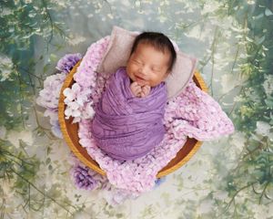 A sleeping newborn wrapped in purple fabric lies in a basket with pink blankets and flowers on a floral backdrop.