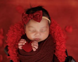 Sleeping newborn wrapped in brown fabric with red bow headband resting on fluffy red blanket holding heart decoration.