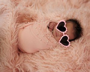 Small infant wearing heart-shaped sunglasses rests on soft pink furry blanket.