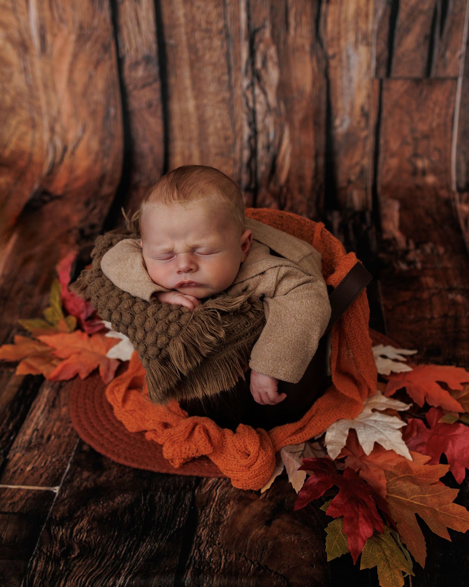 A sleeping newborn nestled among rustic autumn leaves and wooden textures in a warm brown setting.