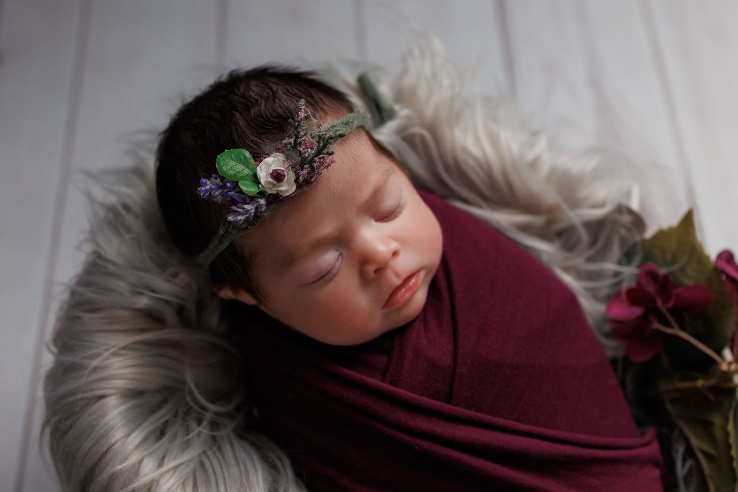 A newborn baby sleeping peacefully during a professional photography session wearing maroon wrap and floral headpiece.