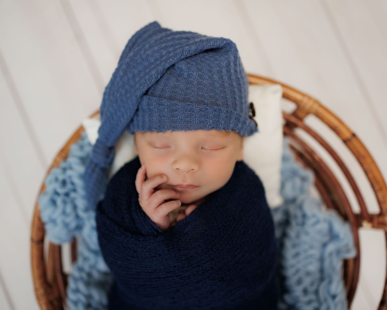 A sleeping newborn wearing a blue knit cap lies swaddled in navy fabric in a wicker basket.