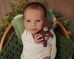 Newborn baby wrapped in white swaddle holding brown knitted teddy bear on green blanket with foliage backdrop.